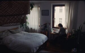 A medium wide shot of a sleeping room and a woman sitting at the table by the window writing. Shot on Zeiss High Speeds.