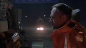 Profile shot of a construction worker wearing an orange vest poorly lit by car lights from the back and construction lights. The triangular shaped lens flares are typical for the Super Speed S16 Zeiss lenses.
