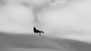 Black and white shot of a woman standing on a dune wearing a black dress. Dust in the background. Shot on a Cooke Varotal zoom lens.