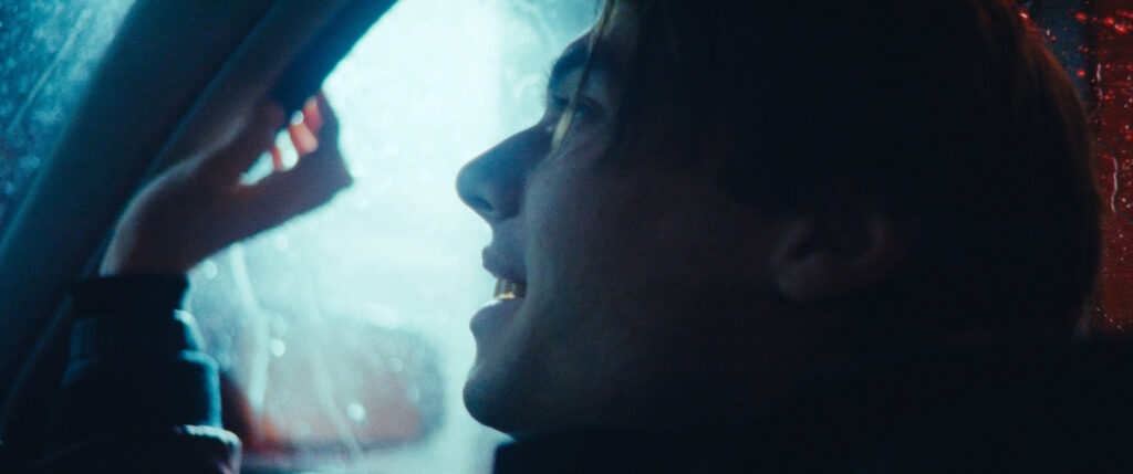 Close up profile shot of a man sitting in a car and smiling. The unsharp background is bright and blue. The raindrops create a bokeh which is typical for Leica lenses.
