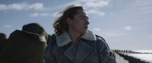 Blond young man in the center of the image infront of breakwaters. Sun is shining, he seems thoughtful. 
Short on LOMO Square Fronts.