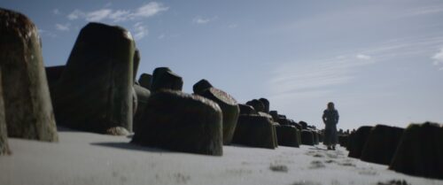 A guy in a coat walking down the beach in between wave breakers. Blue sky with cirrus clouds. 
Short on LOMO Square Fronts.