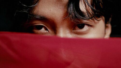 A close up shot of an asian man looking straight into the camera holding a red ribbon in front of his face hiding the nose. Shot with a Leica R cinema prime.