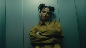 A woman in a yellow shirt is leaning against a metal wall. Top light.