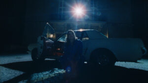 Wide angle silhouette shot of a person kneeling in front of a car lit by a street light which throws a flare into the camera. Captured with a Zeiss Super Speed S16 cinema lens.