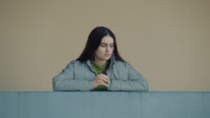 A medium shot of a woman in a blue jacket leaning on a balcony. Timeless feel thanks to the colors and the Cooke Varotal look.