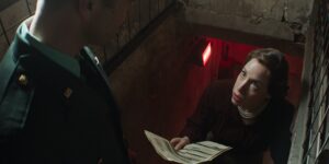 A woman is looking up from a red lit concrete basement facing a soldier. She is holding s document. The Clavius lens is dissorting the picture slightly.