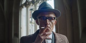 An old man with a hat and glasses is smoking a cigarette. The baroque church window in the background is unsharp due to the open aperture of the Clavius vintage lens.