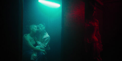 Two women hugging under a green neon light. Another woman standing in the dark red lit room. A Zeiss Standard Speed has been used.