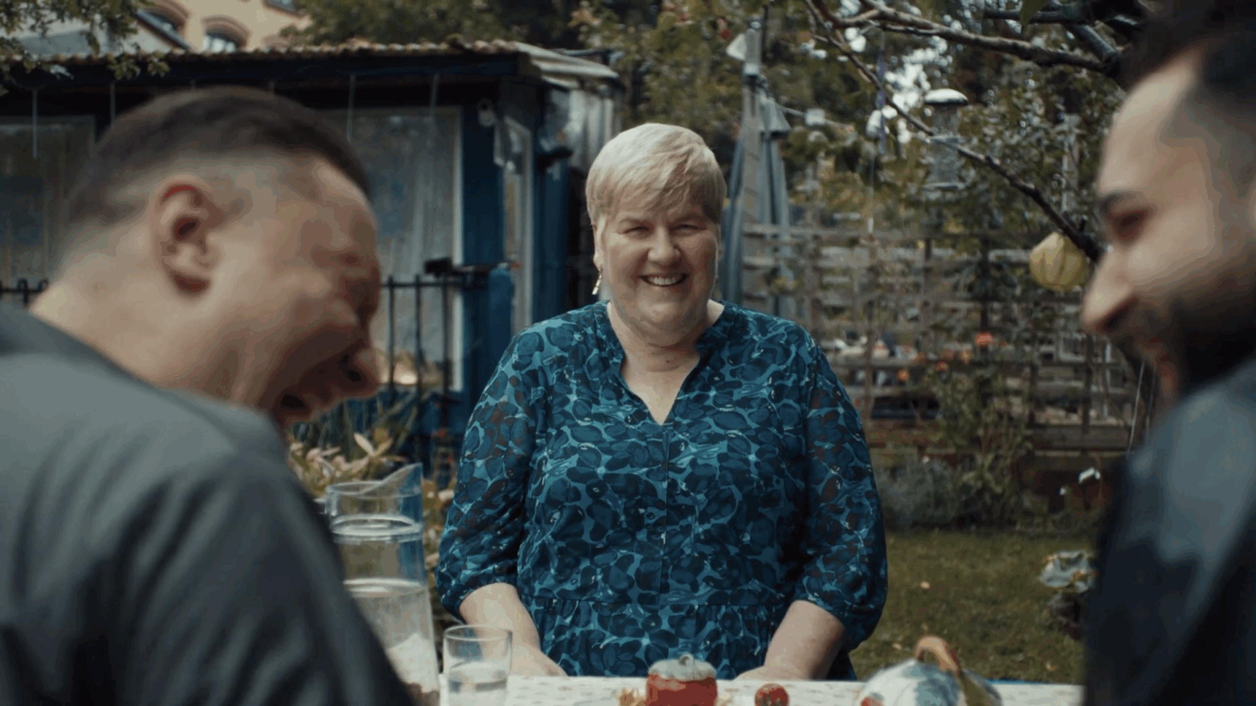 Carmen Goglin sits smiling at a garden table, surrounded by two men in the foreground, in a casual outdoor setting with plants and a small shed in the background.