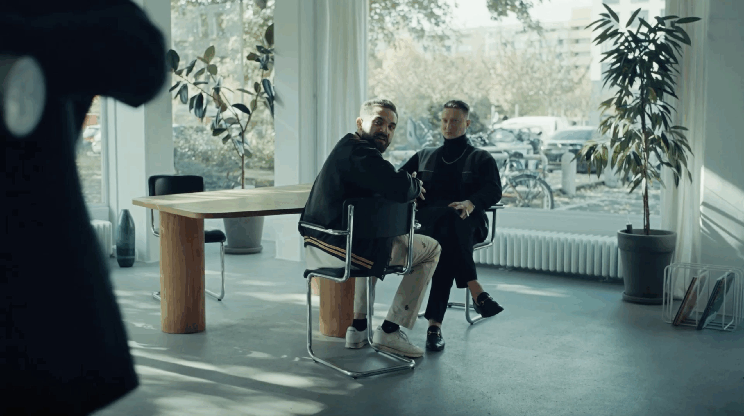 Two men sit in a bright, modern room with large windows and indoor plants, turning their attention toward someone standing off-frame. One man wears a dark jacket and light pants, the other a black outfit with a turtleneck, both seated near a wooden table in a sunlit interior.