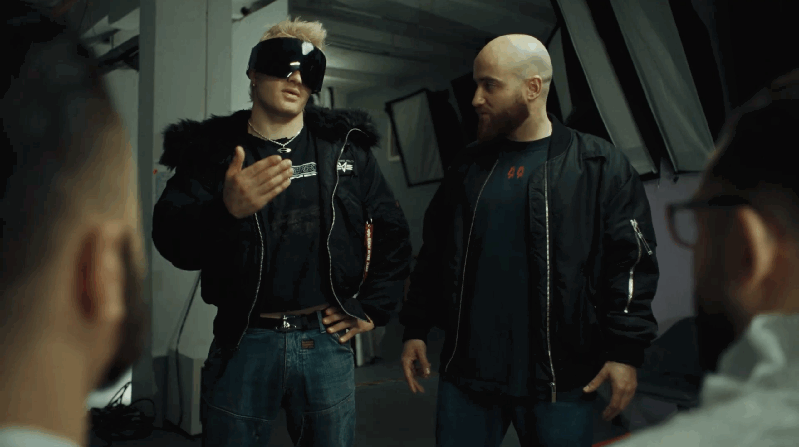 Two men stand one is ski aggu in an industrial-style interior, one wearing large futuristic visor sunglasses and gesturing while speaking, the other listening with a slight smile, as blurred figures frame the foreground.